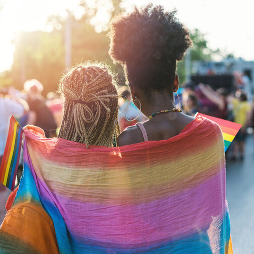 Zwei Frauen stehen Arm in Arm auf einer Veranstaltung im Freien, um die Schulter tragen Sie eine Regenbogenfahne.